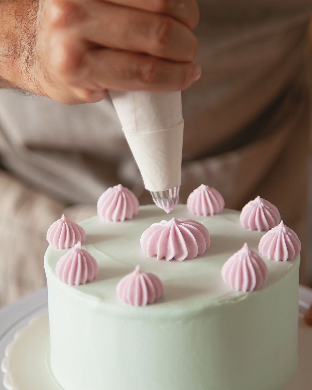 Fotograf&iacute;a de banco con licencia: una mano decora con manga pastelera unos copos de crema rosada sobre un pastel redondo de bet&uacute;n verde claro.